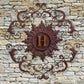 Monogrammed Iron Wall Grille - Monogram Wall Decor | Estate Quality Home Decor | Personalized Wall Decor | Shown with the Monogram "H" | Iron finished in a rustic brown-gold stain with Italian gold 5" monogram shown on stone wall | INSIDE OUT | InsideOutCatalog.com