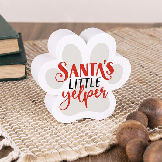 White paw-shaped sign with 'Santa's Little Yelper' text on a textured surface with books and acorns | InsideOutCatalog.com
