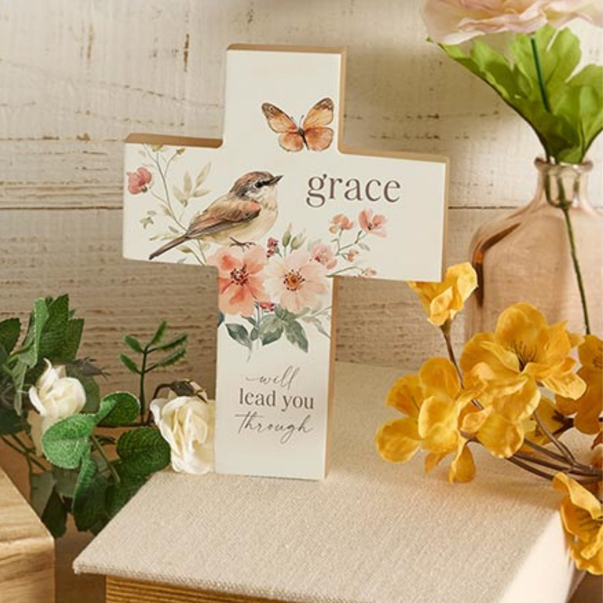 Grace Will Lead You Through Wood Block Table Cross with Butterfly, Bird, & Floral Artwork placed on a surface with flowers and plants | InsideOutCatalog.com