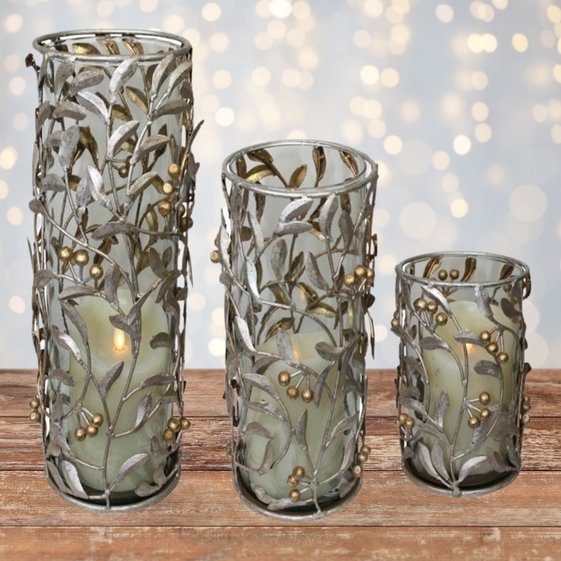 Silver and Gold Iron Hurricanes - Leaf & Berry Accented Iron & Glass Candle Holders - 3 Sizes to Choose From | Iron Candle Holders shown on wood table in front of twinkling lights | Christmas Decor | INSIDE OUT | InsideOutCatalog.com